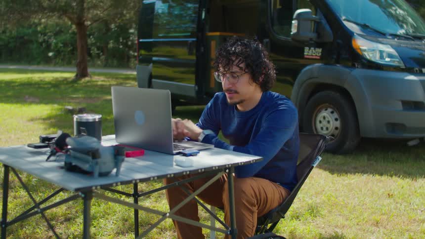 A man sits near a van at a table where he uses a laptop computer