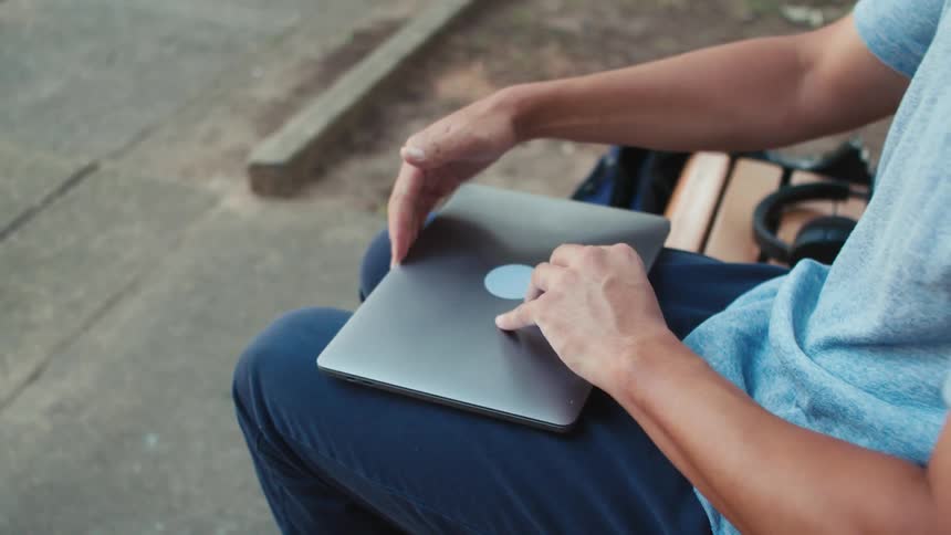 A white male opens a laptop while sitting on a bench outside