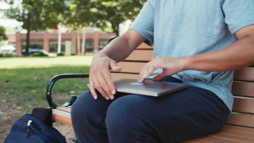 Man sitting on a park bench pulls laptop from his backpack and opens it on his lap