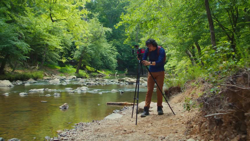 Male photographer sets up tripod for a camera on a riverbank in the woods