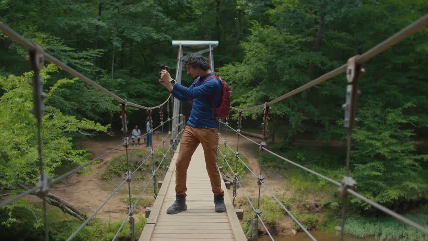 A man on a wood and wire bridge near a forest holding a small camera and microphone