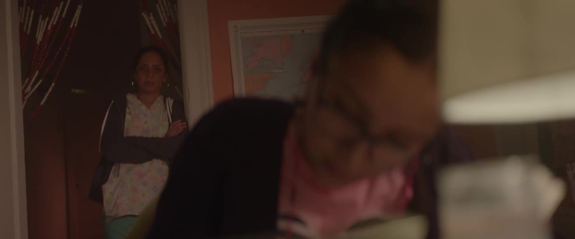 A woman stands in a doorway and watches her daughter do homework