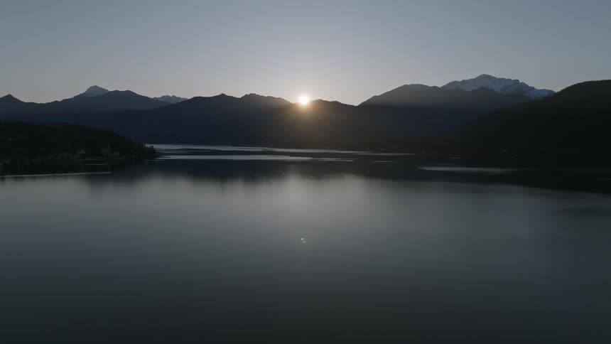A calm lake reflects the sky at dawn with the sun peering over the mountains