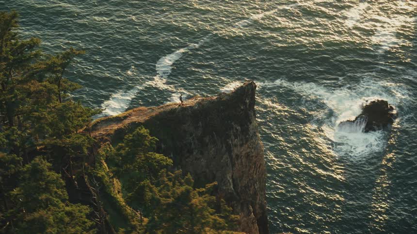 A person walks on a high rocky cliff overlooking the ocean at sunset
