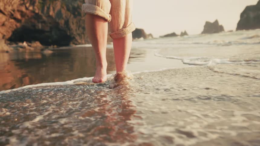 A woman with her pants rolled up to her knee walks through the tide at a beach