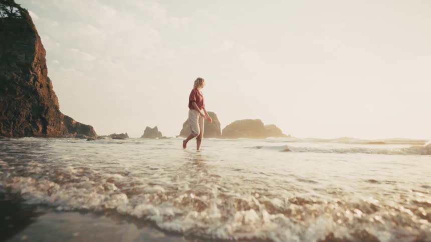 A woman on a beach walking at waters edge with rolled up pants