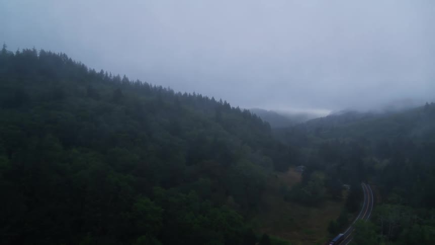 An aerial view of a winding road cutting through a dense forest with low hanging clouds over the hills