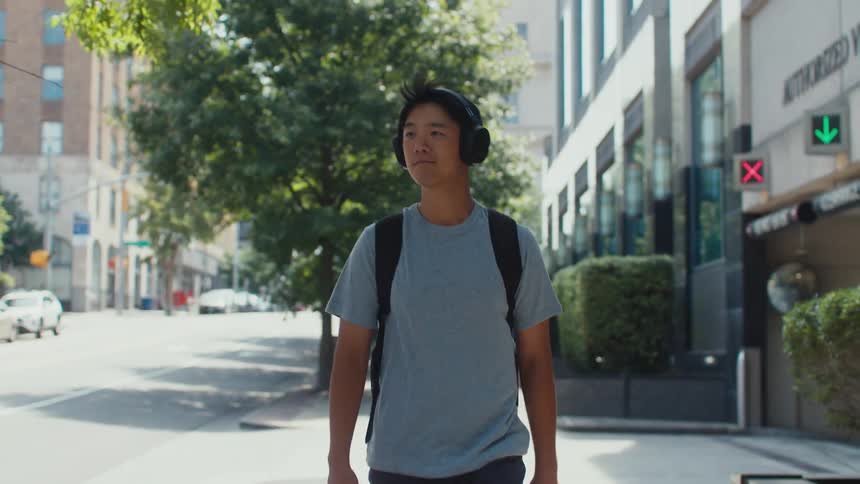 A young man strutting down a city street with his headphones on and a slight smile on his face