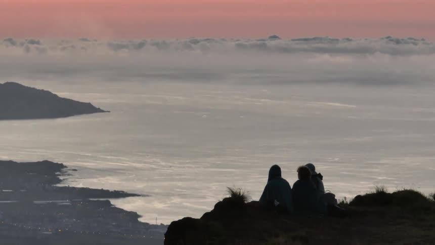 Friends sit on a promontory overlooking a foggy coastal town under a red sky