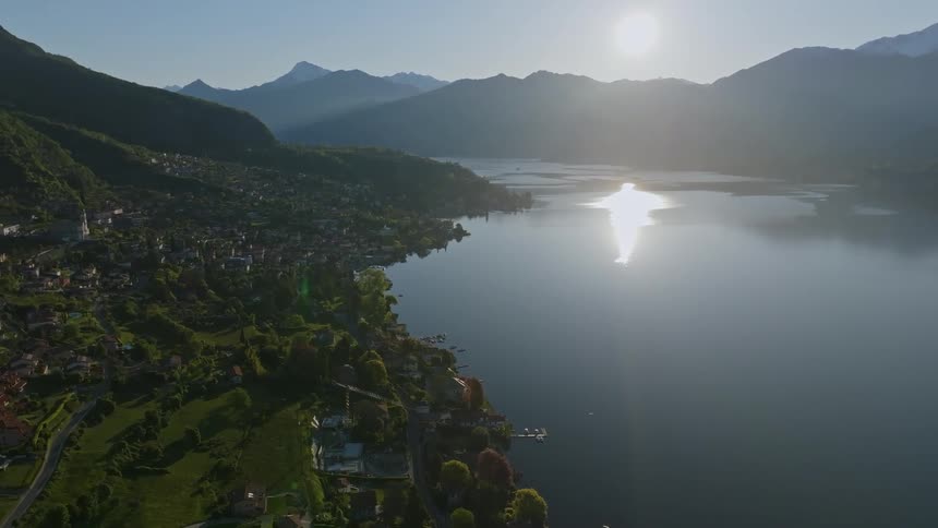 Aerial shot of a natural landscape with a lake surrounded by mountains with a community and the sun on the horizon