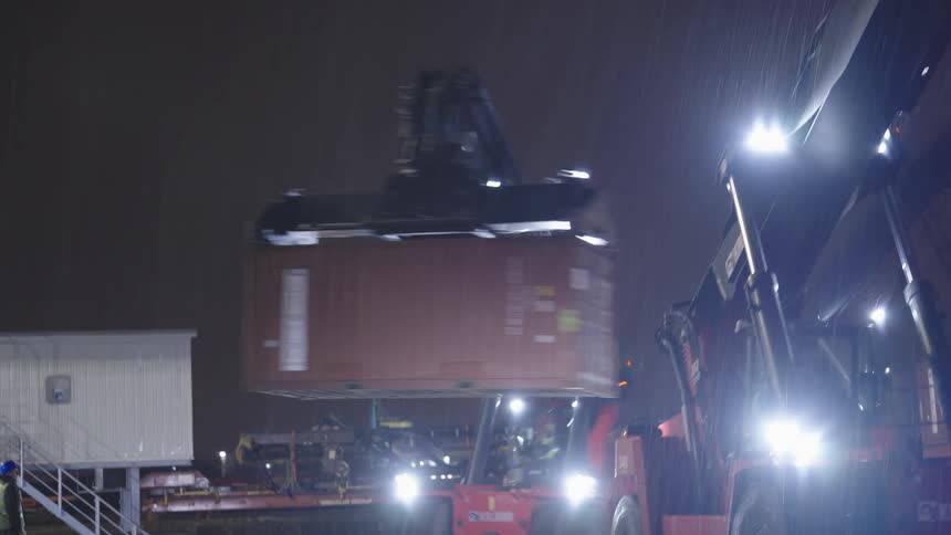 A large container is being lifted by a crane at a shipping yard on a rainy night