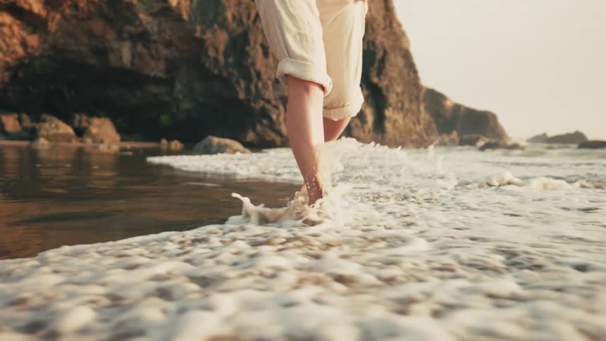 A person walks barefoot on a wet sandy beach her pants rolled up to avoid getting wet