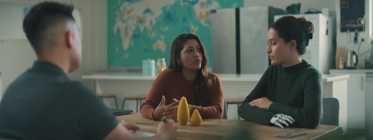 Man sitting at a table with two women one of whom has a robotic hand and they are talking to each other