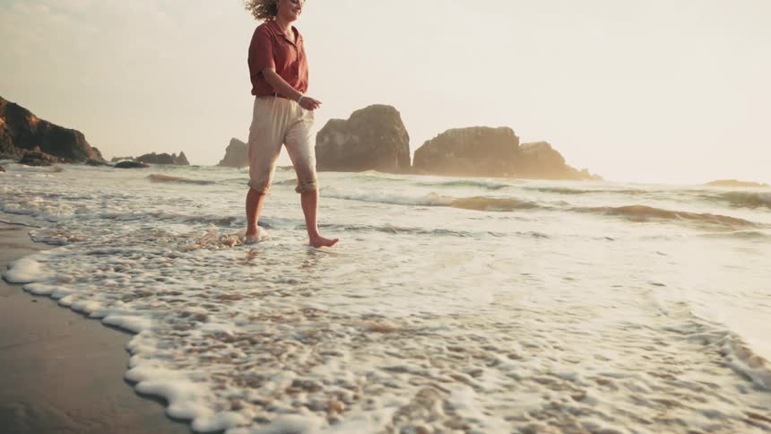 A happy woman walking barefoot on the coast of a beach at sunset