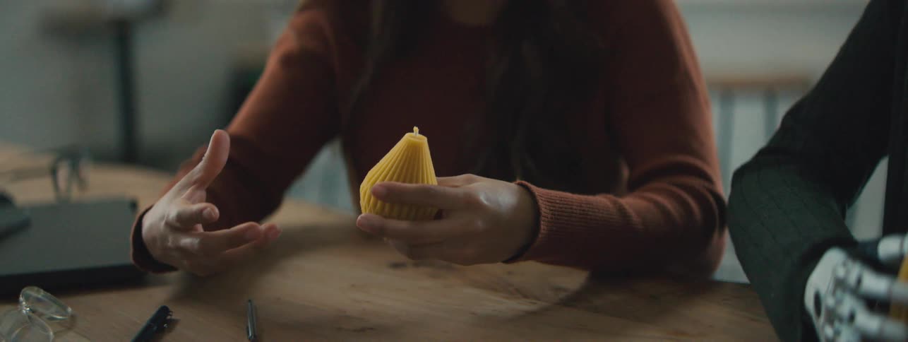 Two women one with a robotic hand and the other human holding abstract yellow objects on the table