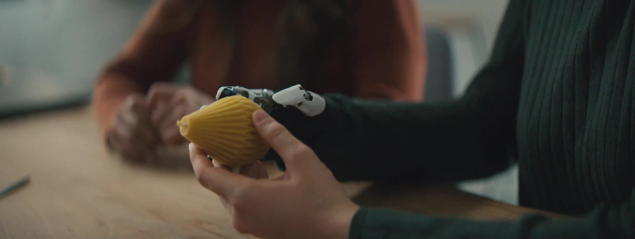 Robotic and human hand holds a yellow object next to a woman at a table