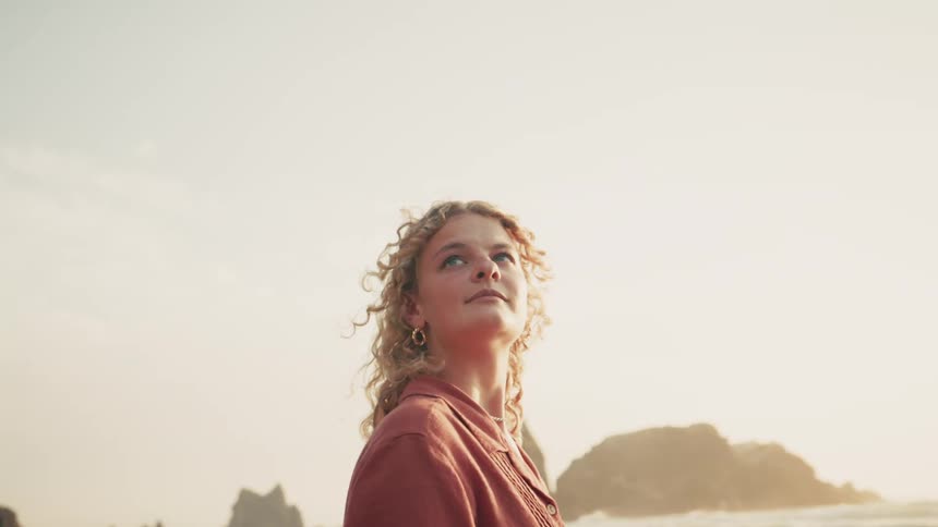 Blonde woman with curly hair on a beach with rocks in the background