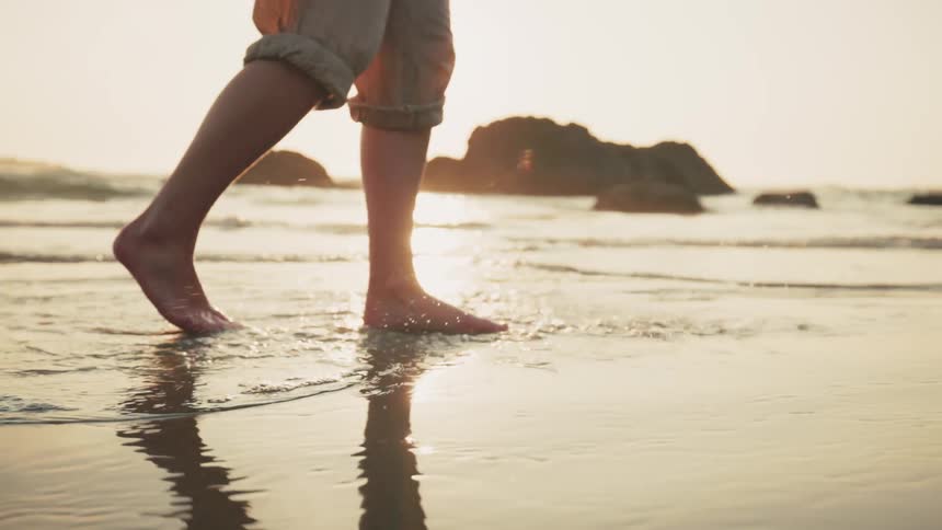 Someone walking on a beach at the waters edge barefoot and with their pants legs rolled up