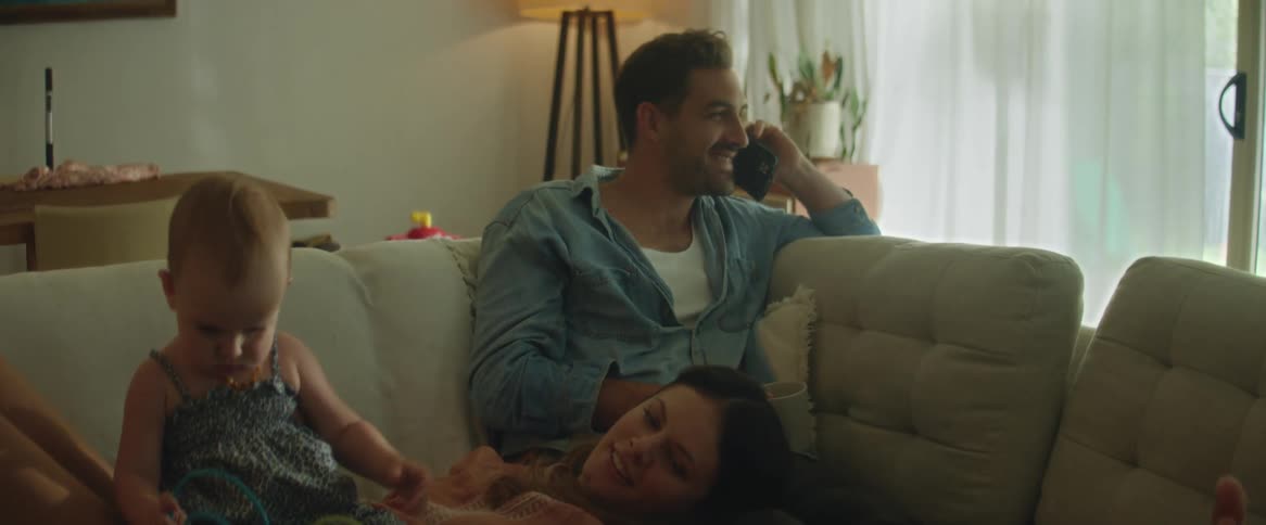 Parents lying on a sofa with the father talking on the phone while the mother plays with their baby daughter