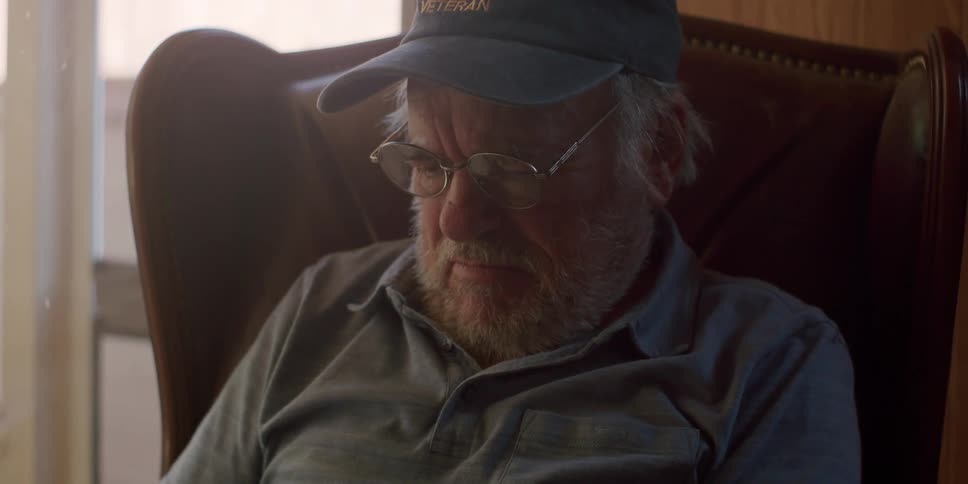 An older man wearing glasses and a veteran cap sits in a leather armchair looking down with a thoughtful expression while using a tablet