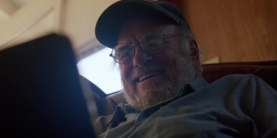 An older man with a beard and glasses smiles and laughs as he looks at a tablet computer seated in a comfortable chair