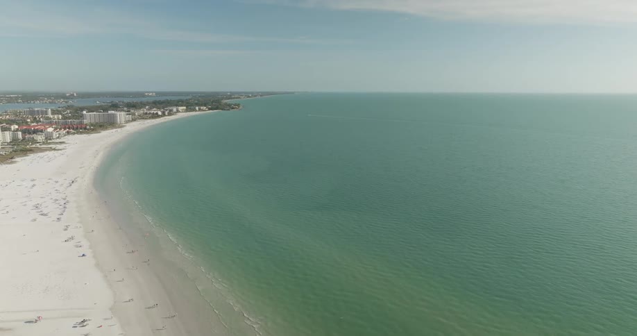 An aerial view captures a long stretch of white sand beach bordered by turquoise waters and a line of buildings in the distance