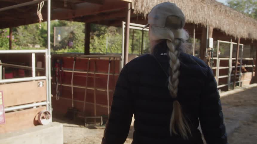A woman with long blonde braids walks towards a row of stables on her way to care for horses