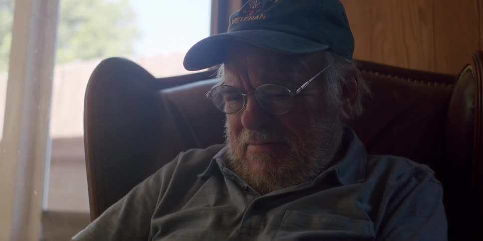 Elderly man with a blue cap glasses and a gray shirt sits in an armchair using a tablet