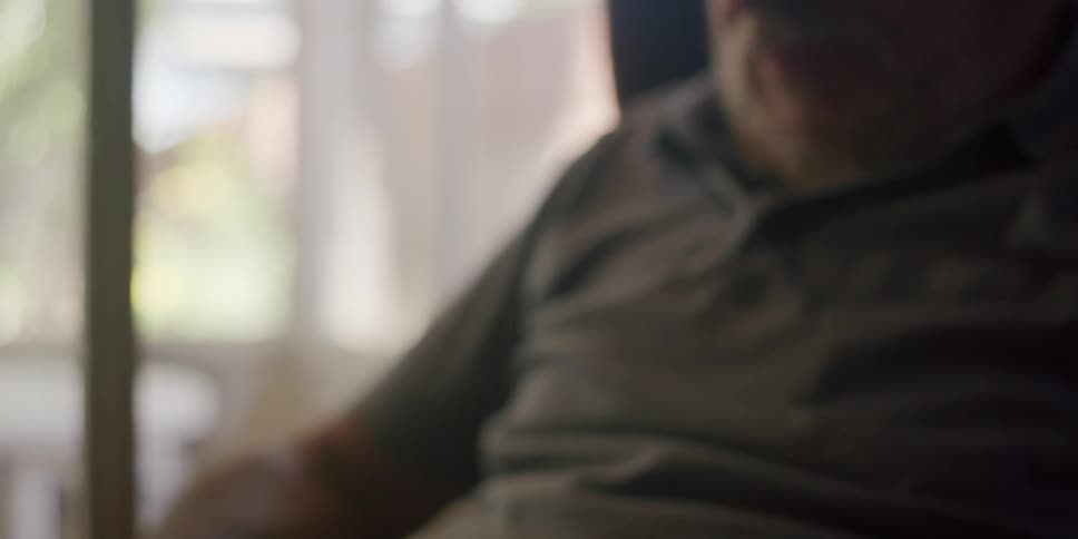 A bearded man wearing a cap and a gray shirt sits in an armchair using a tablet