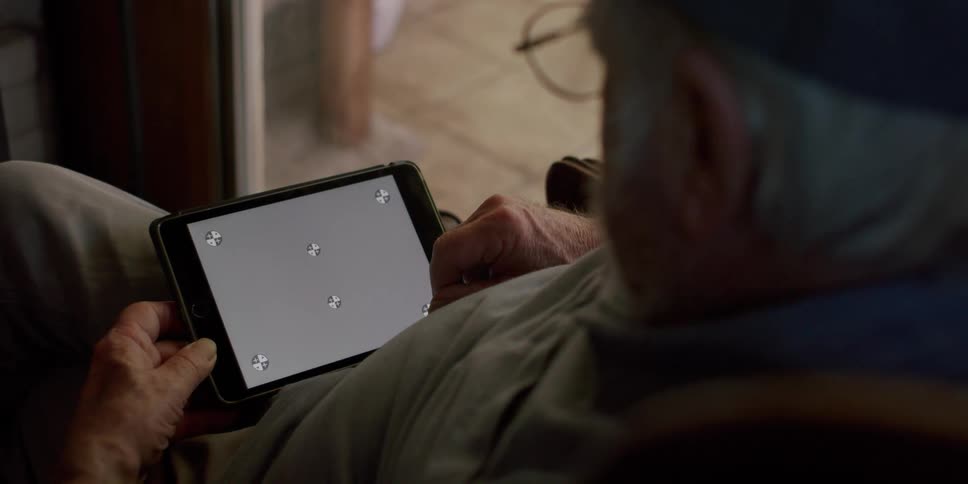 An elderly man plays a video game on a tablet while sitting indoors