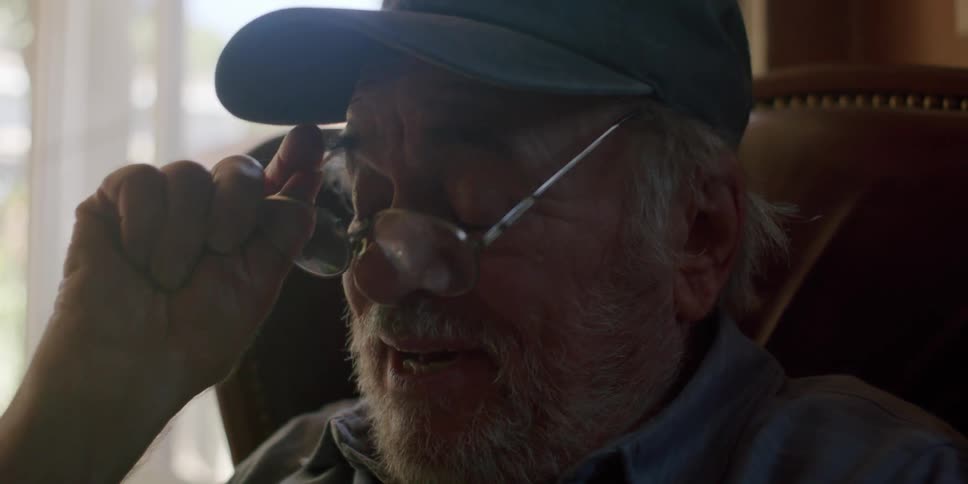 Elderly man sitting in an armchair putting on glasses