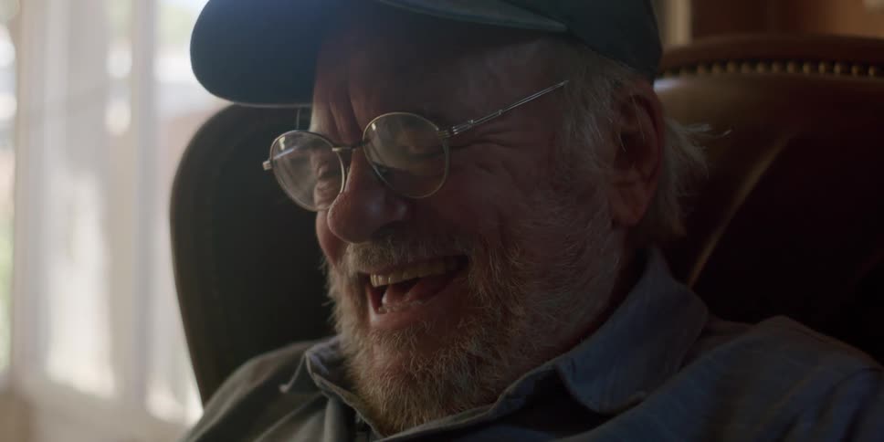 Bearded elderly man wearing a cap and glasses looks down at something and starts laughing