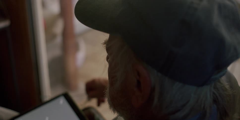 Elderly man sitting in an armchair taking off his glasses and interacting with a tablet