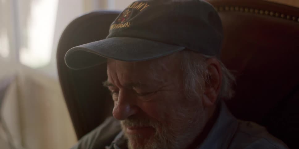 An older man sits in a chair looking thoughtfully before putting on his glasses and using a tablet computer