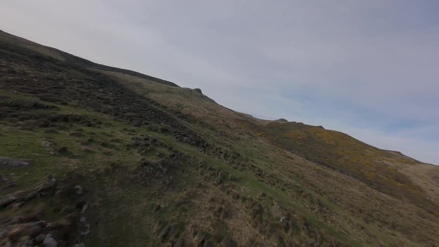 An aerial FPV view shows a vast rolling landscape with sparse vegetation rocky outcrops and hints of yellow wildflowers under a cloudy sky