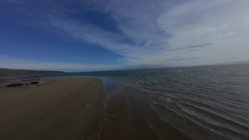A coastline with a long sandy beach meeting the vast blue ocean under a clear sky FPV drone
