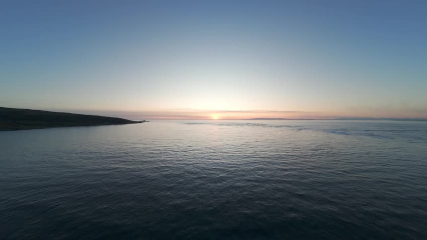 FPV Aerial landscape of the sea with a coast in the background and the sun on the horizon at dusk in moody low light