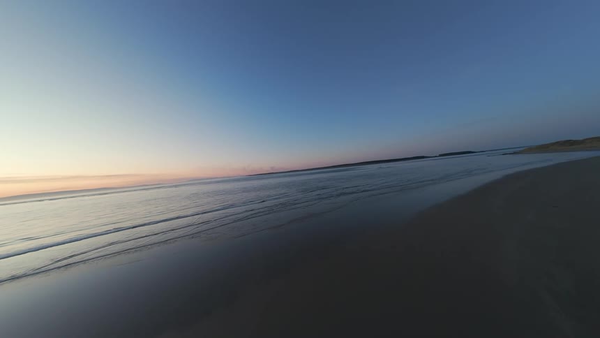 FPV Aerial flight across sand dunes and sandy beach at dawn with low light and sun just dipping below ocean