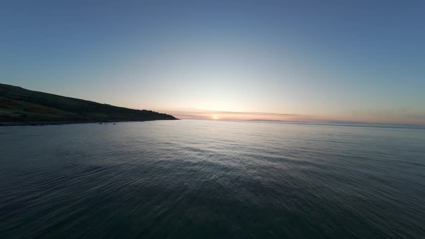 An aerial FPV view of a coastline at dusk with a sandy beach meeting the ocean under a clear blue sky with the sun just dipping
