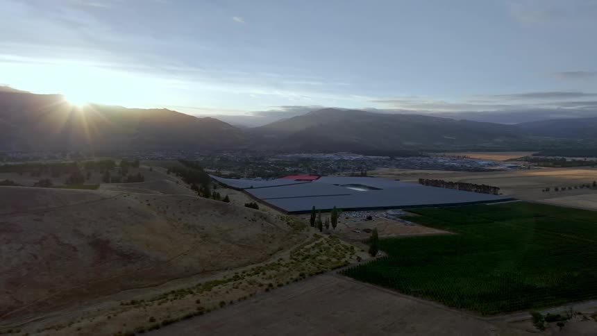 Aerial shot of an agricultural field and a reservoir surrounded by mountains with a town and the sun in the background