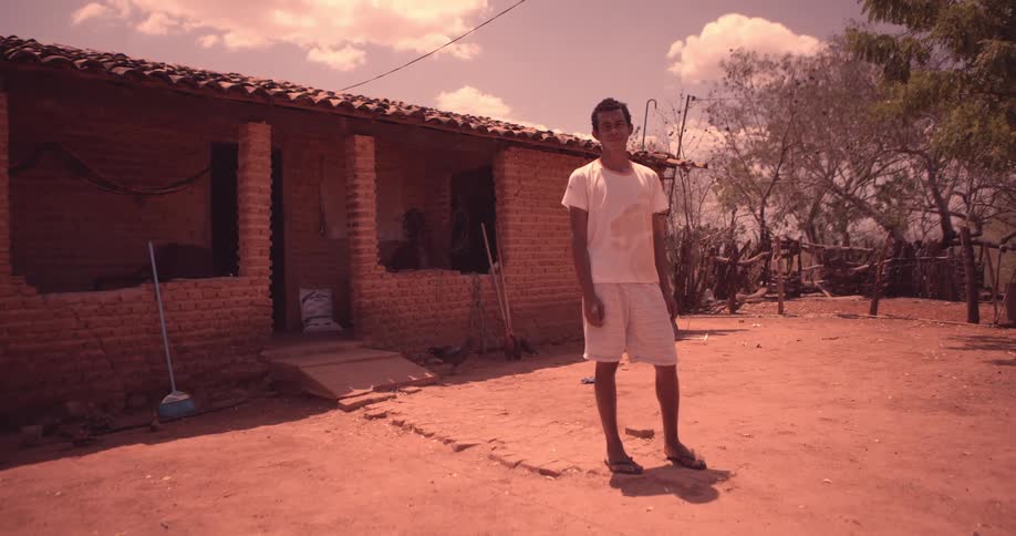 A man in a white shirt and shorts is standing on dry earth and behind him there is a small brick house