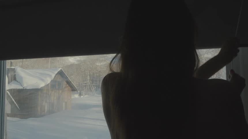 A woman is turned away and is pulling a rope to open the curtain of a glass window and the outside environment is of some houses covered in snow