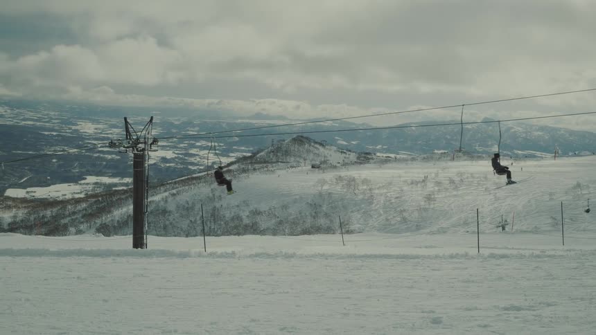 A chairlift with some people sitting being transported to the right in a snowy environment and with some people skiing around