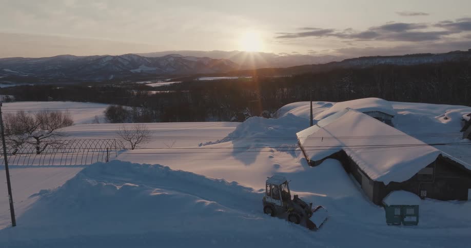 Aerial view of a tractor removing snow from the street and a house next door with mountains and the sun on the horizon