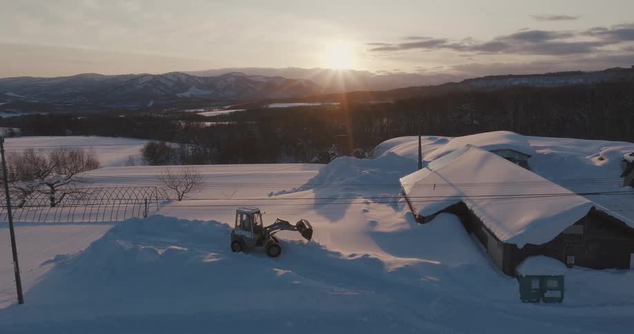 Aerial shot of a tractor clearing snow from a path with a cabin next to it covered in snow and the sun on the horizon