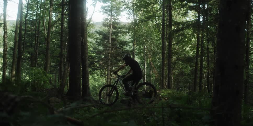 A person standing on a bicycle adjusting his helmet and then starts riding down a hill in a forest