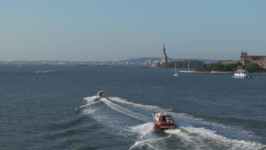 Two small boats sailing on the water and making waves with white foam wherever they go and in the background there is the statue of liberty