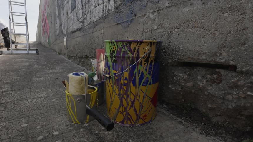 A paint can with splatters of paint on it is sitting on the ground next to a roller and a tray
