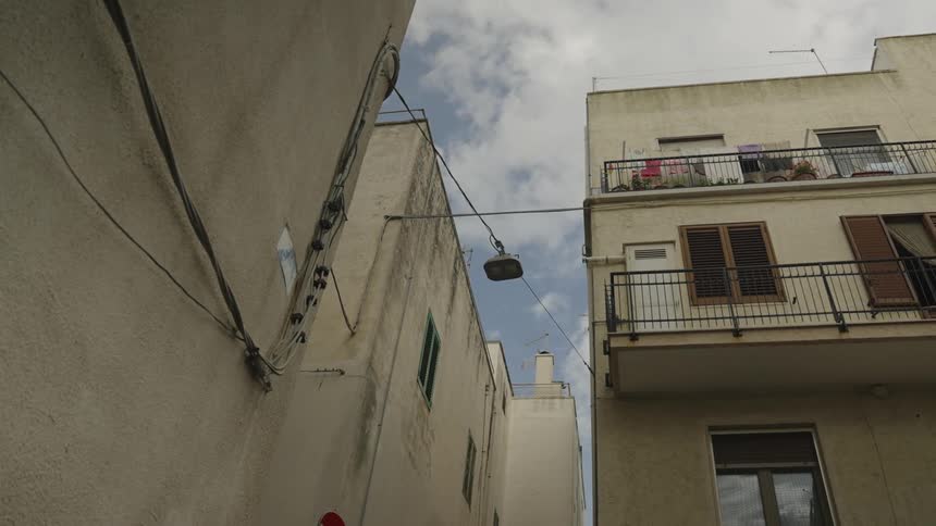 A narrow urban scene with two closely built buildings a suspended streetlight a partially cloudy sky and a balcony with plants and laundry