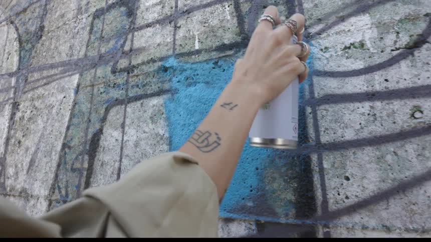 A woman wearing a brown jacket is spraying blue paint on a wall